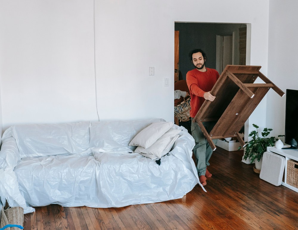 A man carries a wooden table past a sofa wrapped in protective plastic in a bright living room.