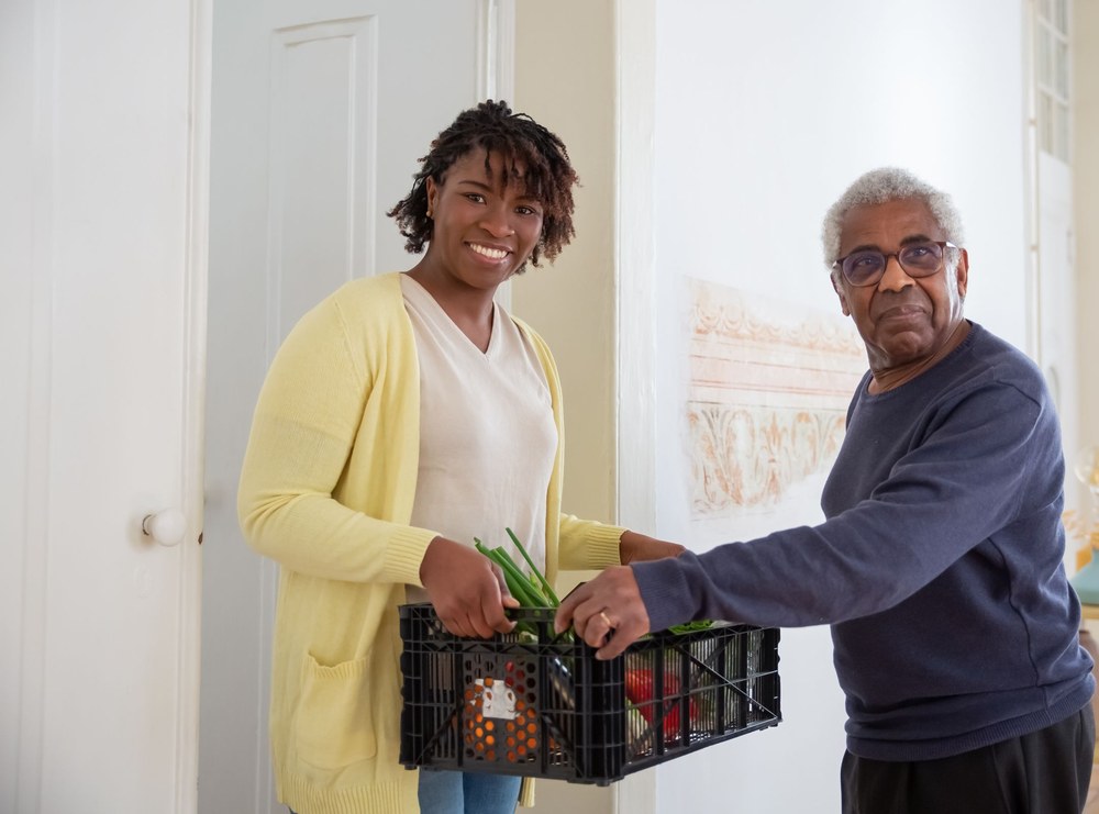 A smiling woman in a yellow cardigan and an older man in glasses stand by an open door, holding a crate of groceries together.