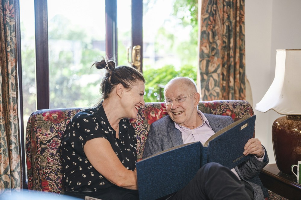 A younger woman and an older man in a suit jacket laugh together while looking through a photo album on a floral sofa.