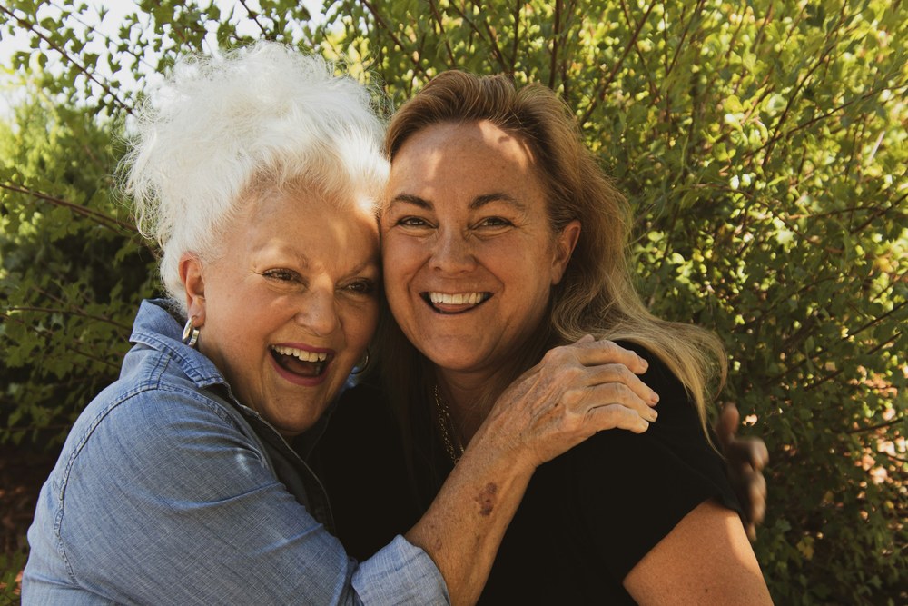 A white haired older woman and a younger woman hug and laugh together outdoors.