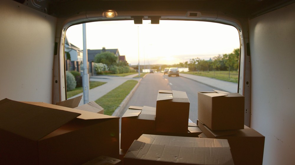 Cardboard boxes loaded in the back of an open van, with a quiet neighborhood street and evening sun beyond.
