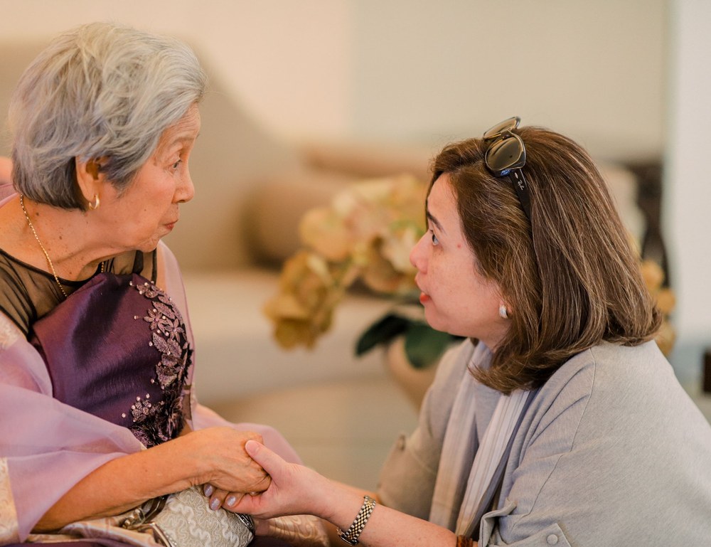 A gray haired older woman in a lavender shawl holds hands with a younger woman who kneels beside her, listening closely.