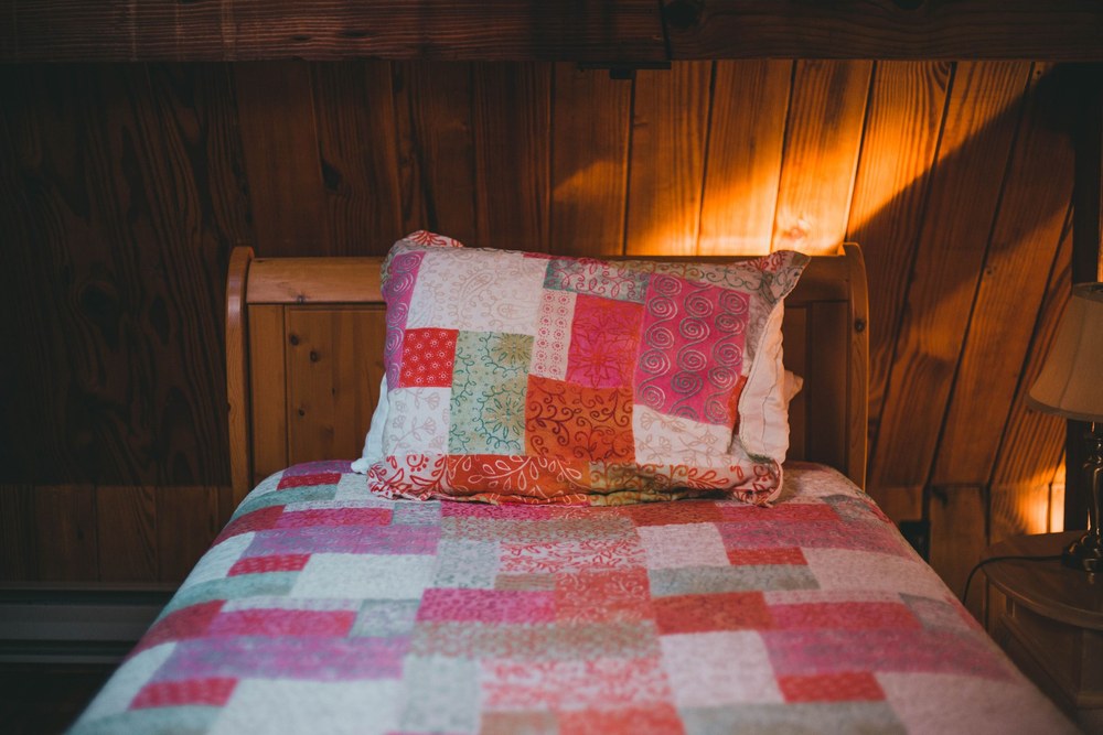 A neatly made bed with a colorful patchwork quilt and pillow in a warm, wood paneled bedroom lit by a lamp.