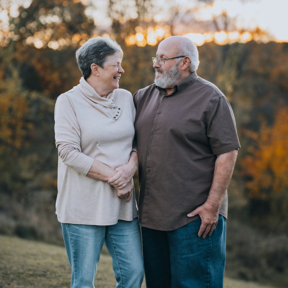 An older couple stands close together and smiles at each other in a grassy field, with autumn trees behind them.