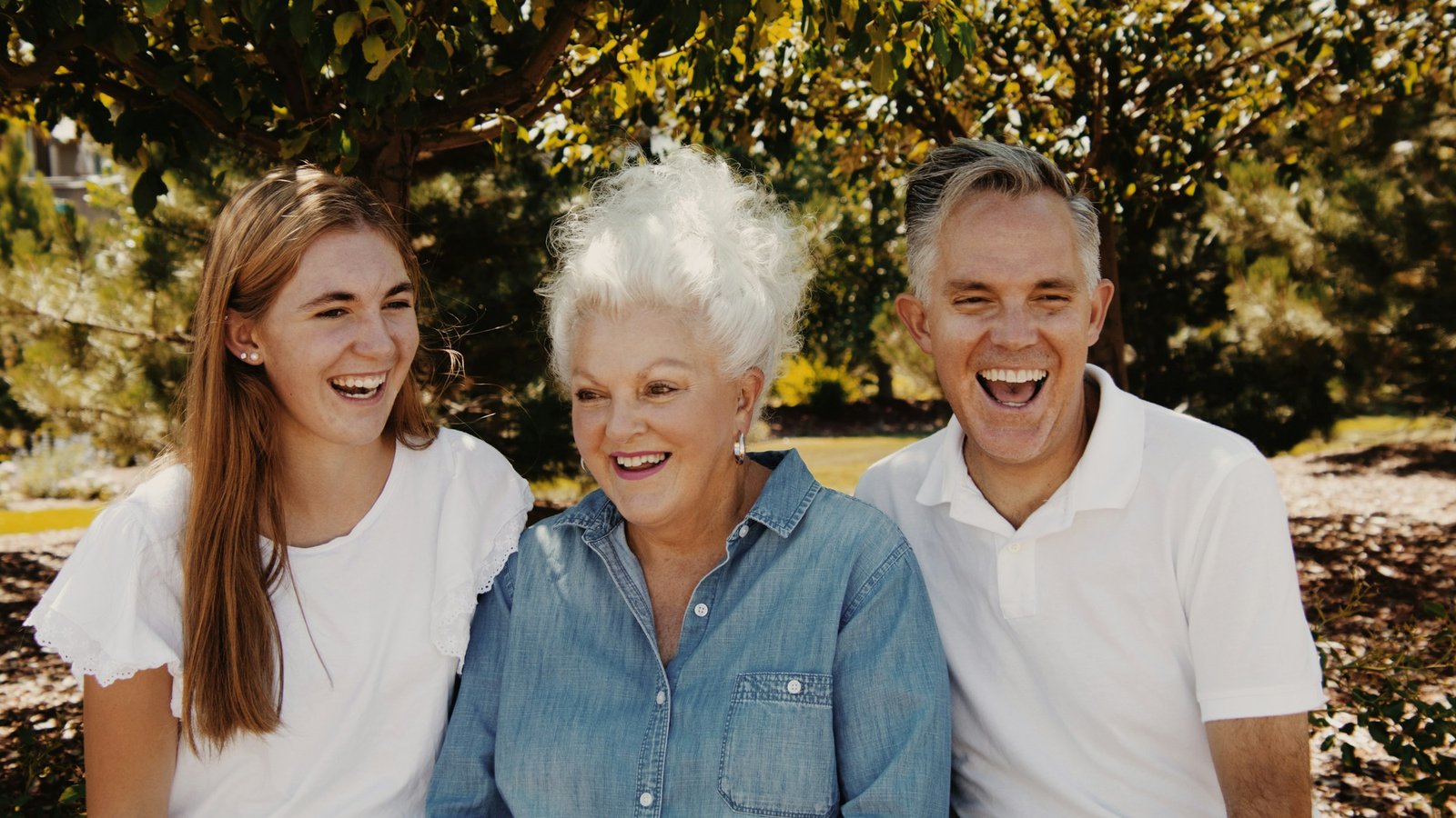 A smiling older woman with white hair sits outdoors between a laughing young woman and a smiling middle aged man.