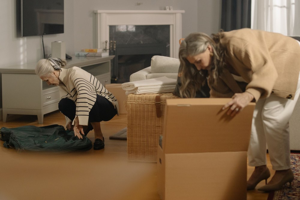 Two older women pack belongings into cardboard boxes in a sunny living room.
