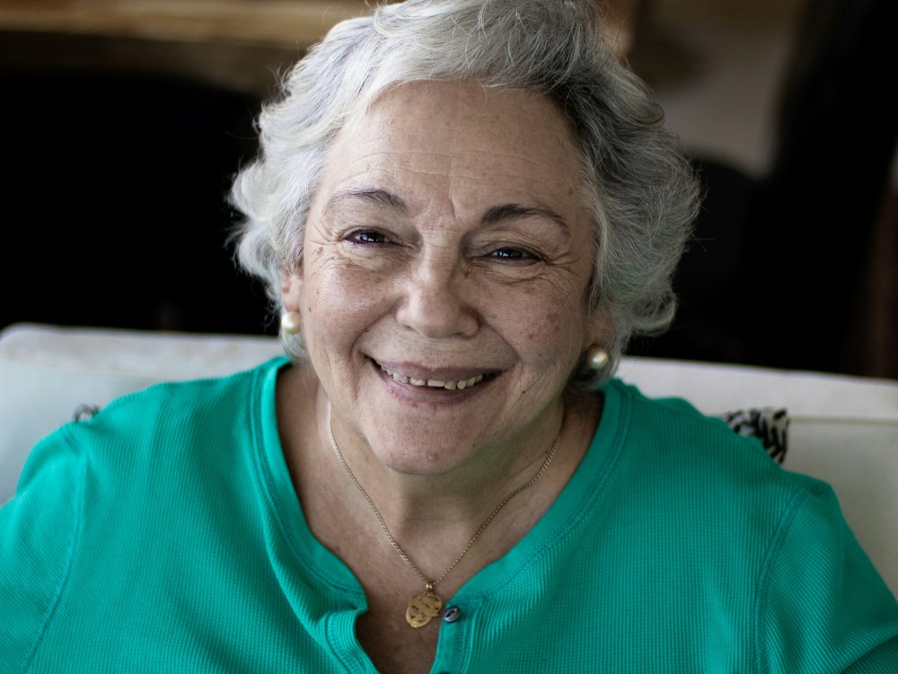 A smiling older woman with short gray hair, wearing a teal top, sits in a living room.