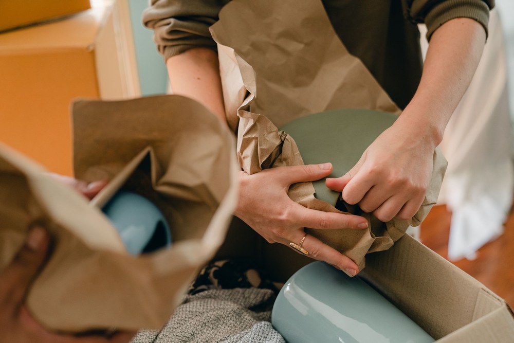 Close up of hands wrapping a ceramic dish in brown packing paper over an open moving box.